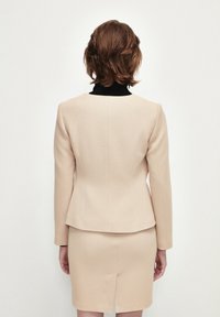 Beige skräddarsydd blazer med strukturerade axlar, texturerad tyg och en svart innerkrage, ihop med en matchande kjol med en slits bak.