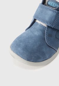 Blaue Wildlederschuhe mit einer strukturierten Oberfläche, ausgestattet mit einem Klettverschluss und einer hellgrauen Gummisohle. Nahaufnahme des Zehenbereichs.