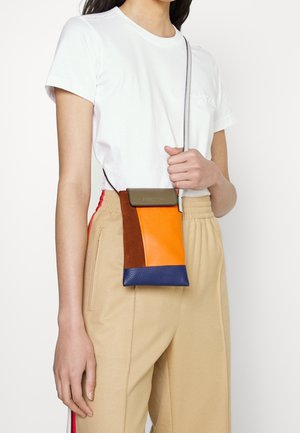 Vrouw draagt beige broek en wit T-shirt, houdt een kleine schoudertas met kleurvlakken in oranje, bruin, blauw en olijfgroen vast.