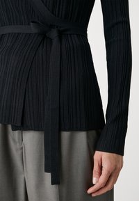 Svart ribbad stickad topp med långa ärmar, med en omslagdesign och knytdekor vid midjan, i kombination med grå byxor.