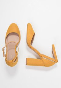 Żółte zamszowe buty na wysokim obcasie z paskiem na kostkę, w szpic, oraz masywnym obcasem. Charakteryzują się gładką teksturą i minimalistycznym wzornictwem.