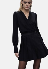 Abito nero con design avvolgente, maniche lunghe e gonna plissettata. Realizzato in tessuto morbido con vita aderente e dettaglio in cintura di raso.