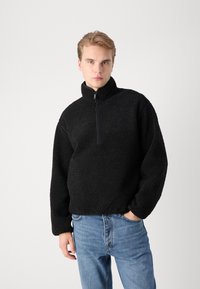 Zwarte fleece trui met hoge kraag, halve ritssluiting en een gestructureerd oppervlak. Gecombineerd met blauwe jeans van denim, met een relaxte pasvorm.