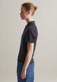Zwarte poloshirt met een gestructureerd patroon en korte mouwen, gedragen met hoge denim broeken, gezien vanaf de zijkant.