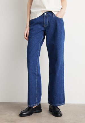 Personne portant un jean bleu droit, des mocassins noirs et un haut blanc, debout avec une main dans la poche sur un fond uni.