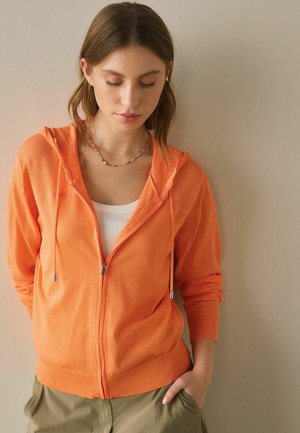 Junge Frau mit braunen Haaren, die einen orangen Kapuzenpullover mit Reißverschluss, ein weißes Oberteil, eine bunte Perlenkette und grüne Hosen trägt, mit einer Hand in der Tasche.