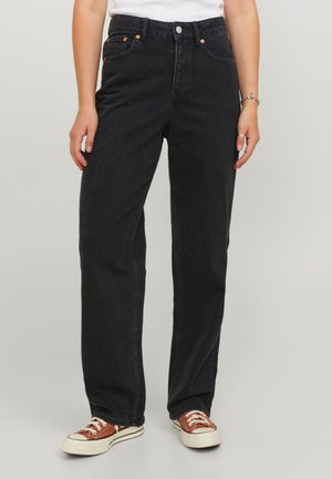 Personne portant un jean noir taille haute à jambes droites et des baskets basses marron avec des lacets blancs, debout sur un fond clair uni.