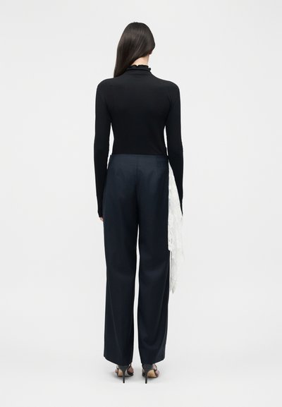 Γυναίκα όρθια με την πλάτη γυρισμένη, φοράει μαύρο μακρυμάνικο τοπ και ναυτικό μπλε παντελόνι φαρδιάς γραμμής με λευκή δαντέλα στη δεξιά πλευρά.