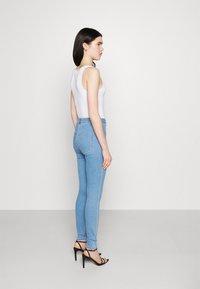 Jeans skinny em denim azul claro com uma cintura alta, combinados com uma parte de cima branca sem mangas e sandálias de salto alto pretas com tiras, vistos de lado.