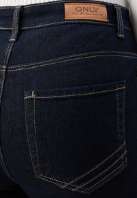 Tamno plavi traperice od denima s kožnom etiketom na kojoj piše "ONLY." Imaju vidljive šavove i jednu stražnju džep s suptilnim detaljima.