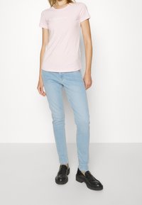 Světle růžové bavlněné tričko s kulatým výstřihem a krátkým rukávem, se bílým logem na hrudi. Nosené s světle modrými slim jeans a černými botami.