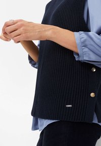 Marineblaues geripptes Strickwestenoberteil mit goldenen Druckknöpfen an der Seite, über einem hellblauen Hemd mit umgeschlagenen Ärmeln getragen. Strukturierte Oberfläche, lockere Passform.