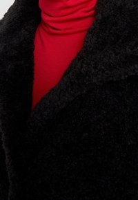 Svart fluffig kappa med strukturerad yta, utrustad med en vid krage, lagd över en röd polotröja. Mjuk look med kontrasterande färger.
