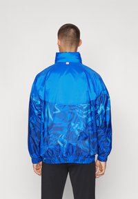 Giacca a vento blu con un design testurizzato in diverse tonalità di blu, polsini elasticizzati e un piccolo logo sul colletto.