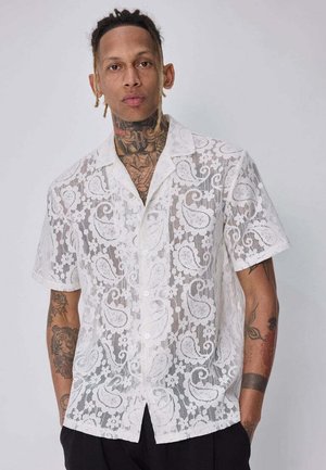 Homme avec des tatouages porte une chemise blanche en dentelle à manches courtes avec des motifs cachemire et un pantalon noir, debout devant un fond uni.