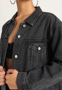 Černá denimová bunda s opranou texturou, zapínáním na knoflíky a dvěma náprsními kapsami. Má klasický límec a stříbrné kovové doplňky.