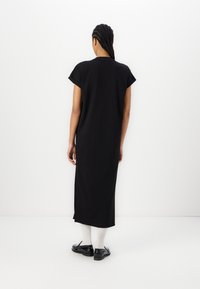 Personne avec des cheveux tressés portant une robe midi noire à manches courtes, des chaussettes blanches et des mocassins noirs, debout face à un mur blanc.