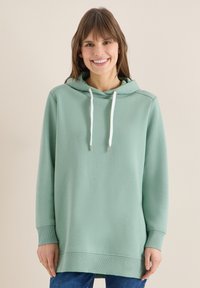 Femme souriante portant un sweat à capuche oversize vert clair avec des cordons blancs et un jean bleu, sur un fond beige uni.
