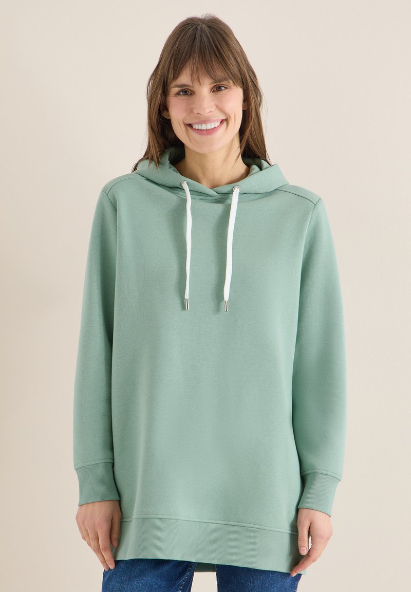 Femme souriante portant un sweat à capuche oversize vert clair avec des cordons blancs et un jean bleu, sur un fond beige uni.