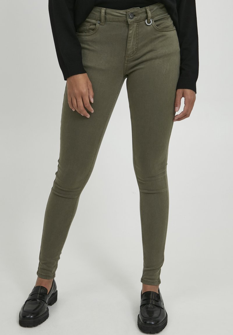Jeans skinny verde oliva con tasche anteriori, dettagli di cucitura discreti e un accento in anello metallico sulla vita. Abbinati a mocassini neri.