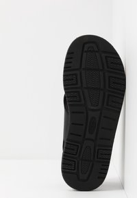 Semelle de chaussure noire avec motifs texturés de la bande de roulement et lignes d'adhérence, debout verticalement sur un fond blanc d'angle.