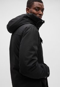 Chaqueta negra con capucha, de textura suave, con cierre de cremallera, capucha ajustable con cordón y detalles de bolsillos laterales.