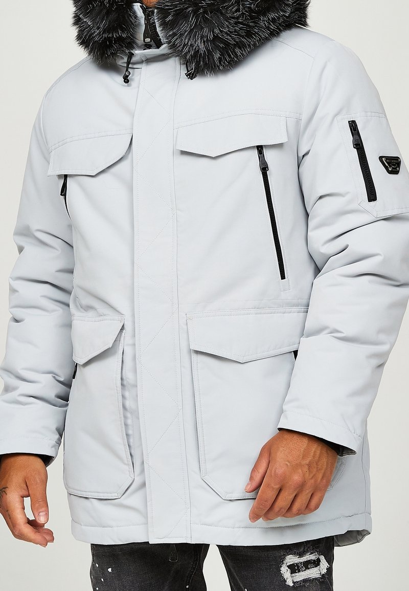 Homme portant un manteau d'hiver gris clair avec une capuche bordée de fourrure, plusieurs poches à rabat et zippées, et un patch sur la manche gauche.