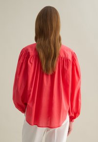 Blusa di cotone rossa con maniche a palloncino, vestibilità comoda e polsini con bottoni. Il retro presenta una cucitura a yoke e una texture fluida.