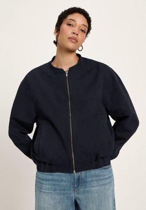 Frau mit kurzen lockigen Haaren, die goldene Creolen, eine marineblaue Jacke mit Reißverschluss und hellblaue Jeans vor einem einfarbigen Hintergrund trägt.