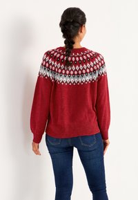 Roter gestrickter Pullover mit einem gemusterten Passe in Grau, Schwarz und Weiß. Lange Ballonärmel und ein gerippter Saum. Getragen mit blauen Jeans.