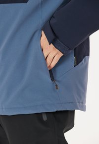 Wasserabweisende blaue Jacke mit navyfarbenen Akzenten, ausgestattet mit einer Reißverschlusstasche und strukturiertem Ärmelabschluss. Der Stoff hat eine glatte Oberfläche.