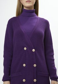 Gilet en tricot violet avec un décolleté en V profond, un design à double boutonnage, deux poches avant et des boutons décoratifs à finition dorée.