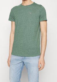 Mężczyzna ubrany w dopasowaną zieloną koszulkę typu crewneck z małym logo na piersi oraz jasnoniebieskie dżinsy, stojący z rozluźnionymi rękami.