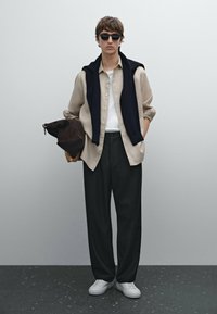Beige Hemd über einem weißen T-Shirt, schwarze Hose, marineblauer Pullover lässig über die Schultern gelegt, braune Tasche in einer Hand, weiße Sneaker, Sonnenbrille.