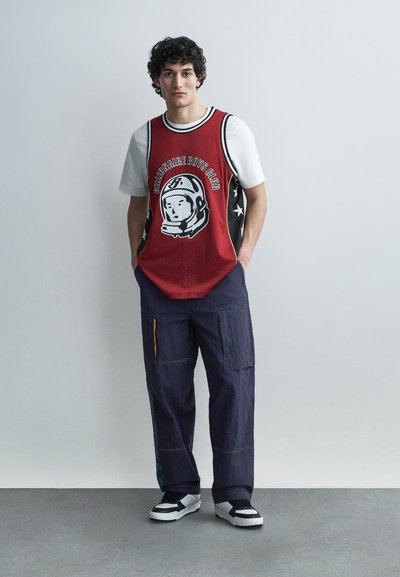 Νεαρός άνδρας φορώντας λευκό μπλουζάκι κάτω από κόκκινο αμάνικο κορυφή "Billionaire Boys Club", ναυτικό cargo παντελόνι και μαύρα-άσπρα αθλητικά, στέκεται με τα χέρια στις τσέπες.