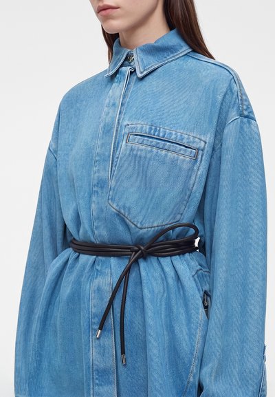 Femme portant une robe veste en denim bleu clair avec un col, une poche poitrine et une fine ceinture noire nouée à la taille.