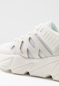 White athletic sneaker with mesh fabric and synthetic overlays, featuring cut-out designs, textured accents, and a chunky sole.