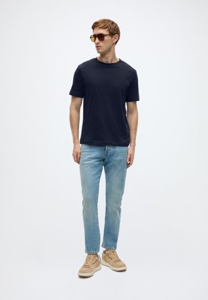 Jūras zila T-krekls ar apaļu kaklu, gaiši zili džinsi un gaiši brūnas sporta kurpes. Modelis valkā brūnas toņa saulesbrilles, stāvot pret baltu fonu.