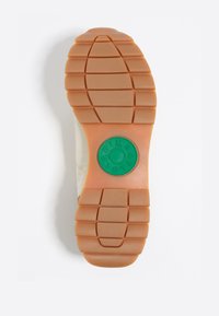 Suela de zapatilla de goma, con una base en tono marrón claro y patrones de tracción texturizados. Un logo circular central en verde aporta un detalle de marca.