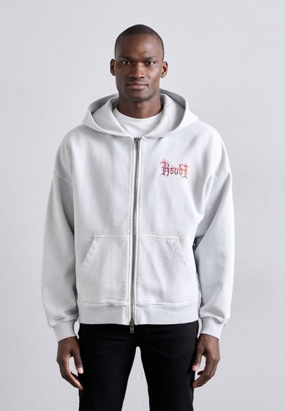 Grå zip-up hoodie med fickor framtill och huva med dragsnöre; har röd grafisk text på bröstet. Bärs över en vit skjorta och svarta byxor.