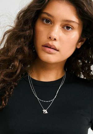 Collares de plata en capas con una cadena retorcida y un colgante que presenta un pequeño diseño, llevados sobre una blusa negra ajustada. Cabello suave y ondulado visible.