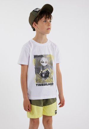 T-shirt en coton blanc avec un design graphique comprenant des éléments noirs, gris et jaunes, associé à un short olive et jaune et une casquette olive.