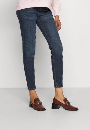 Femme portant un jean slim bleu foncé et des mocassins marron brillants à talons blocs, debout sur un sol gris contre un mur blanc.