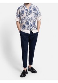 Man in een beige overhemd met korte mouwen en een blauw bloemenpatroon, marineblauwe broek, zwarte loafers, staand met zijn handen in de zakken.