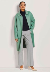 Groene wollen jas met een kraag, grote zakken aan de voorkant, gecombineerd met grijze gemusterde broeken en beige hoge hakken.