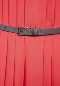 Dettaglio di un abito plissettato rosso, caratterizzato da una cintura in tessuto grigio scuro con fibbia in metallo, che mette in mostra una texture liscia e linee pulite.