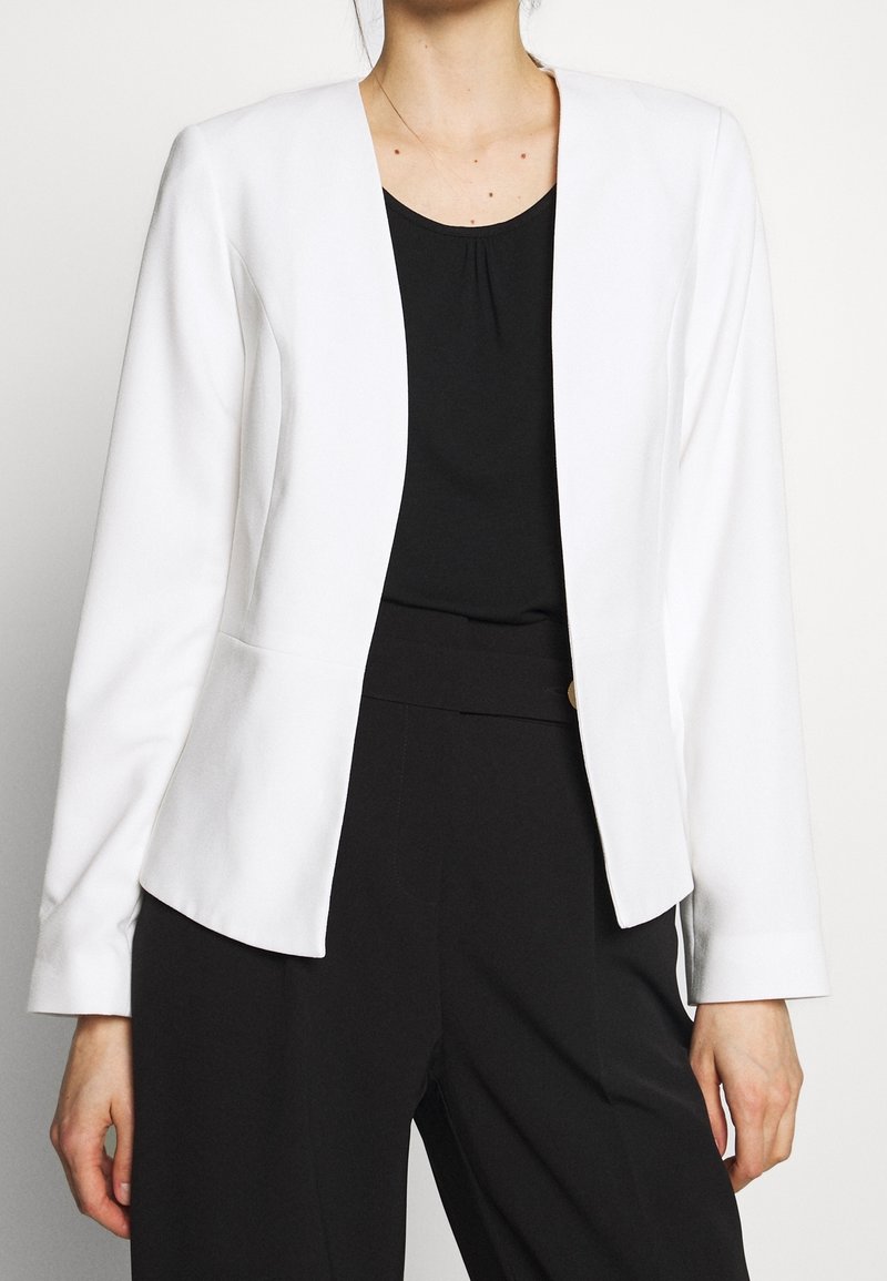 Femme portant un blazer blanc ouvert sur le devant, par-dessus un haut noir et un pantalon noir taille haute, debout devant un fond uni.