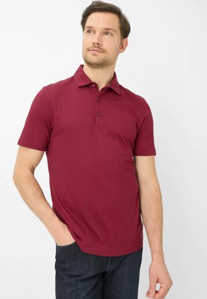 Uomo che indossa una polo bordeaux a maniche corte e jeans scuri, in piedi con una mano in tasca, guarda leggermente verso l’alto a sinistra.