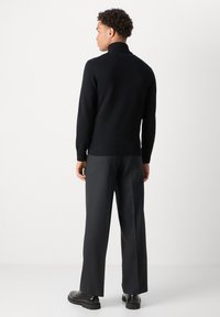 Hombre de espaldas que lleva un jersey negro de cuello alto, pantalones anchos de color gris oscuro y zapatos negros sobre un fondo blanco liso.