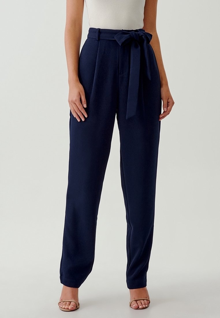 Femme portant un pantalon taille haute bleu marine avec une ceinture en tissu nouée en nœud, associé à un haut sans manches blanc et des sandales à bout ouvert.